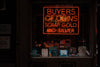 An orange-red neon sign, spelling the words ‘buyers of coins, scrap gold and silver’ hangs in a shop window; upright books on baseball, The Beatles and other topics are arranged below it.